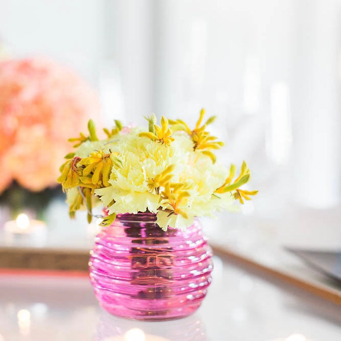 Small purple clear vase with yellow and white flowers on table with peach flowers blurred in background