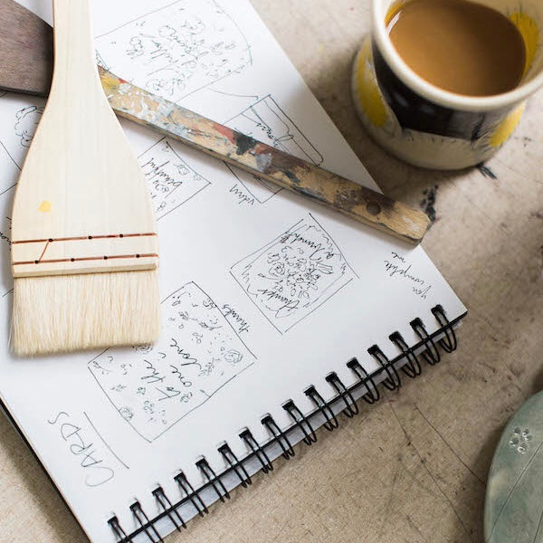sketch book with wooden paintbrushes laid on top and a cup of coffee to its left