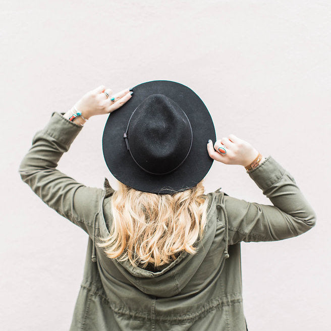 Back image of woman in olive jacket, long blonde hair, black hat and hands grabbing either side of hat