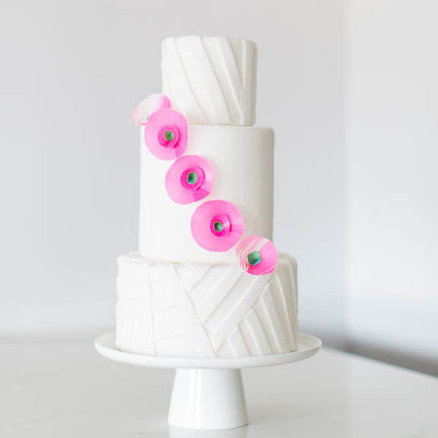 All white 3 tier cake with 4 pink flowers diagonally placed across middle tier on white cake stand